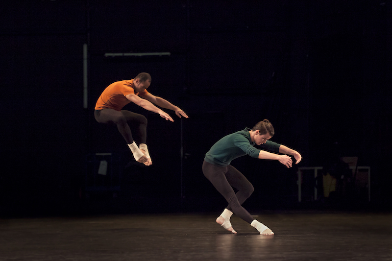 One dancer contracts his torso and pulls his torso back. Another dancer creates a similar a space except that he's jumping.